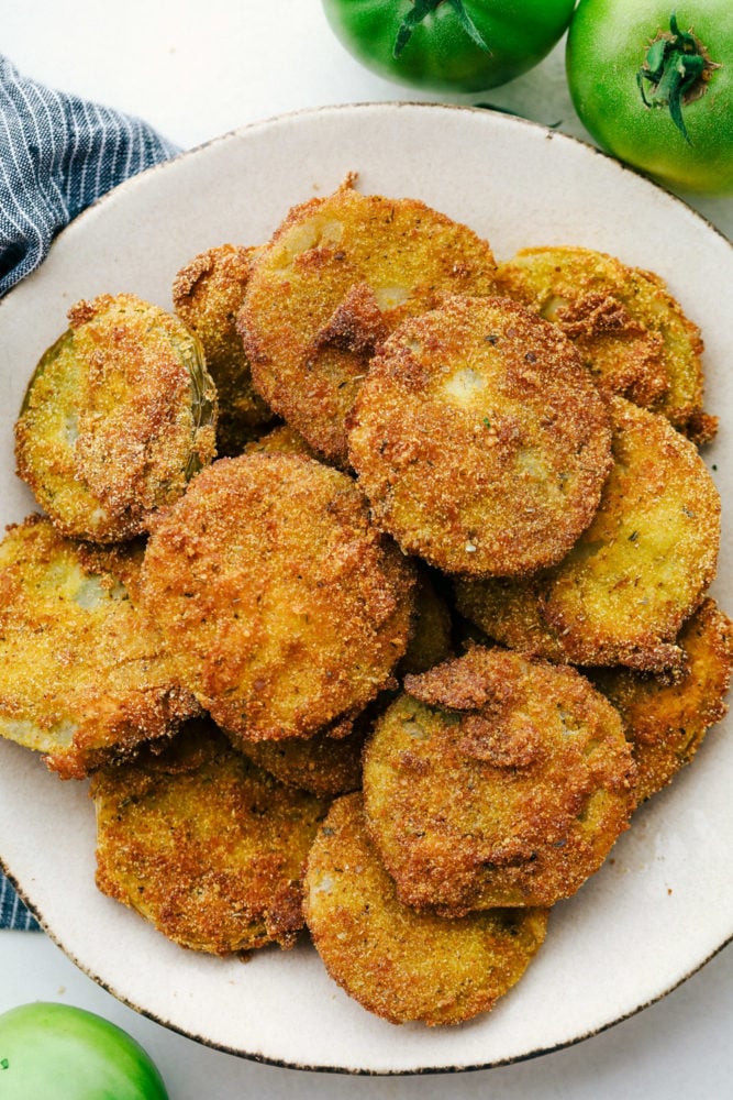Fried Green Tomatoes are extremely popular sliced and fried crunchy bites of tangy green tomatoes. They are totally addictive and easy to make at home! Fried green tomatoes stacked on a white plate.