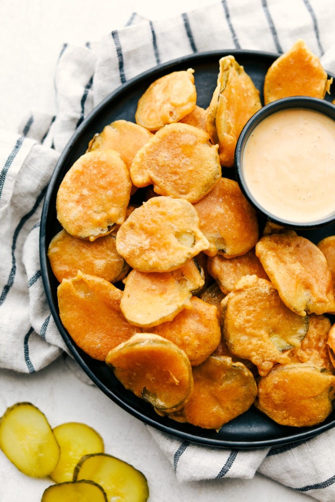 Fried Pickles are tangy dill pickles dipped in a smooth flavorful batter and fried till crispy golden brown. A sure fire winner for any appetizer or snack time! Fried pickles on a black plate with dipping sauce.