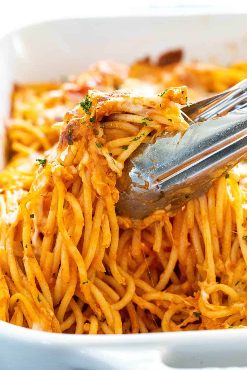 Baked chicken spaghetti is an easy dinner casserole to feed a family. Tender pieces of chicken tossed with tender noodles and a creamy tomato sauce. Tongs lifting a serving of pasta and chicken