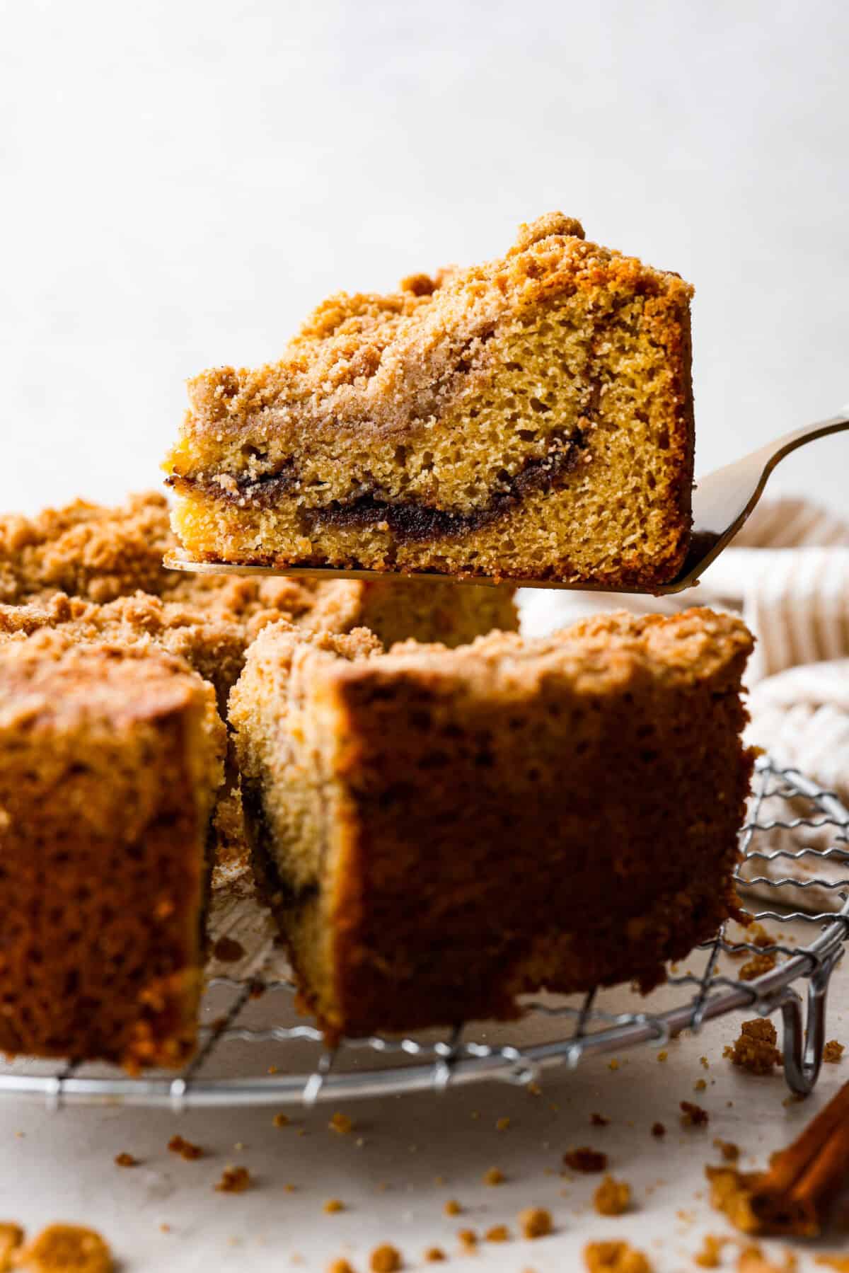This classic cinnamon coffee cake is loaded with decadent cinnamon sugar swirls and topped with crumbly cinnamon streusel. It's the perfect treat to share at any brunch, book club, or dessert for any night of the week. The warm, satisfying aroma will fill your heart and your home! Side shot of a slice of cinnamon coffee cake being lifted off the cooling rack.