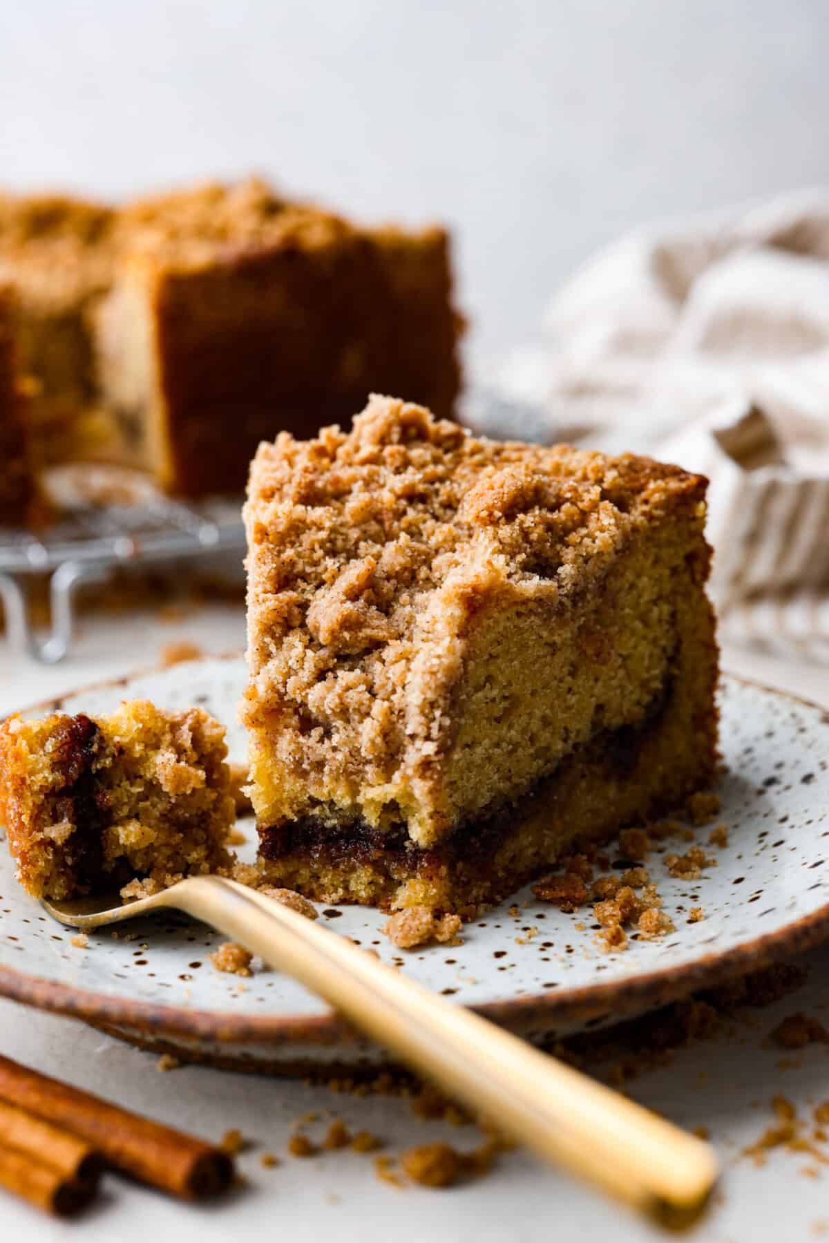 This classic cinnamon coffee cake is loaded with decadent cinnamon sugar swirls and topped with crumbly cinnamon streusel. It's the perfect treat to share at any brunch, book club, or dessert for any night of the week. The warm, satisfying aroma will fill your heart and your home! Close up shot of a slice of cinnamon coffee cake on a plate with a gold fork with a bite on it.