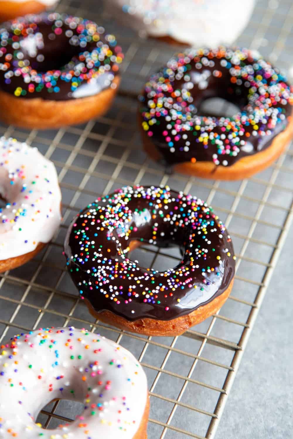Homemade Donuts are easier to make than you might think! And yes, I’m talking about soft and fluffy fried yeasted donuts, dressed in your choice of vanilla or chocolate glaze. Glazed donuts with sprinkles on a wire rack.