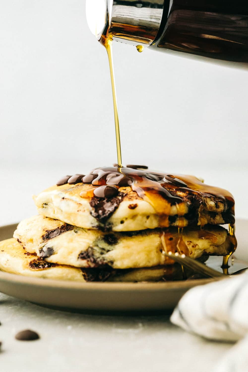 Warm, fluffy, and chocolatey, these Chocolate Chip Pancakes are worth waking up for! Super easy, fast and inexpensive, pancakes are a perfect way to start the morning.ย Syrup pouring on top of chocolate chip panckaes.
