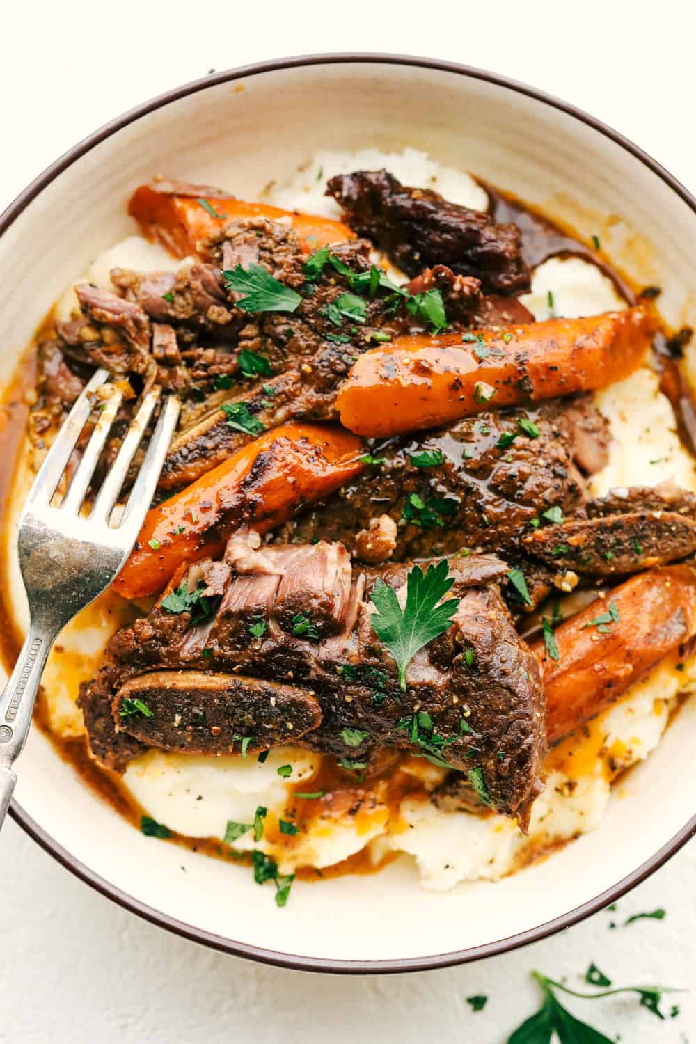 Fall-off-the-bone slow cooker short ribs are a perfectly hearty and savory dinner! This is the best way to cook succulent beef and fork-tender carrots that are emerged in a delicious sauce that is full of flavor. Fork Tender Slow Cooker Short Ribs in a bowl with mashed potatoes and carrots.