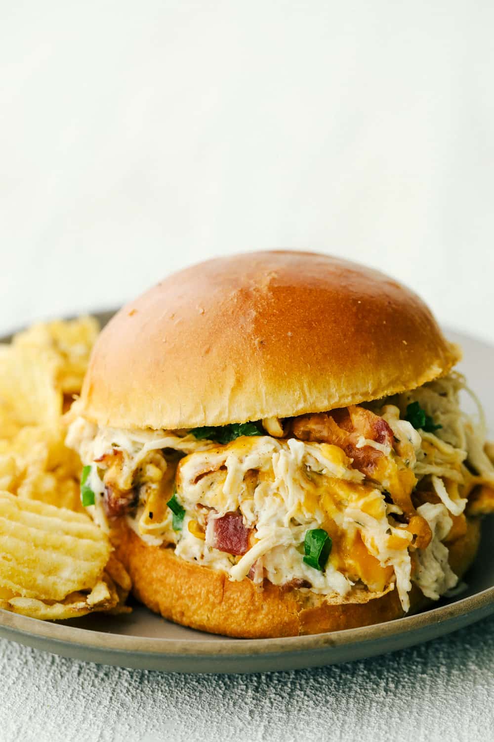 This slow cooker crack chicken (cheddar ranch chicken) is an addicting combination of chicken, cheese, bacon and ranch dressing. Cooking this in the slow cooker takes all the work out of it and makes it a super simple hearty dinner for busy days. Slow cooker ranch chicken served on a bun alongside potato chips.