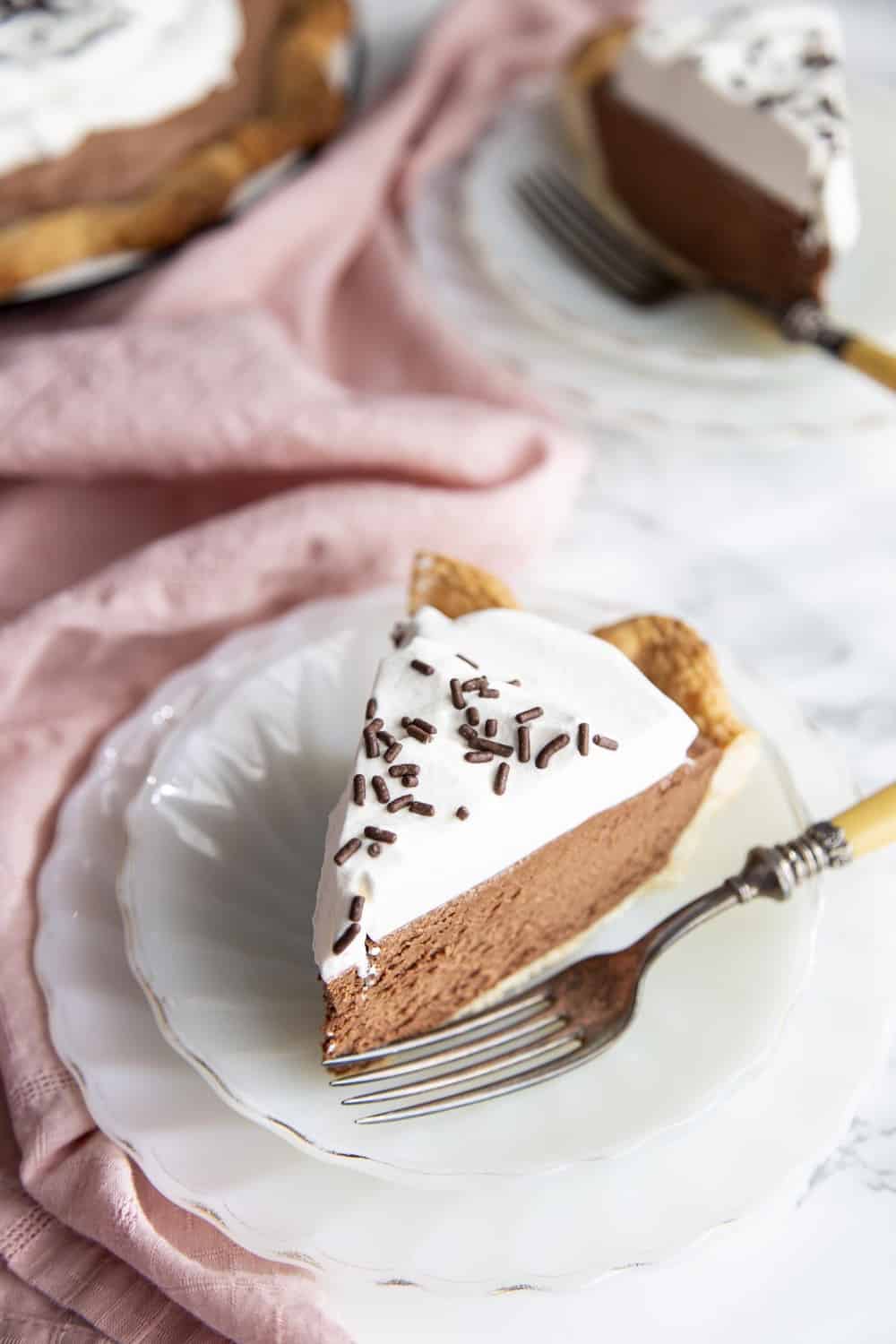 A flakey crust is filled with a decadent, dreamy, creamy, chocolate filling and topped with pillows of cream for this irresistible French Silk Pie. The filling is so smooth and rich, and then encased in a buttery crust. Slice of french silk pie on a plate.