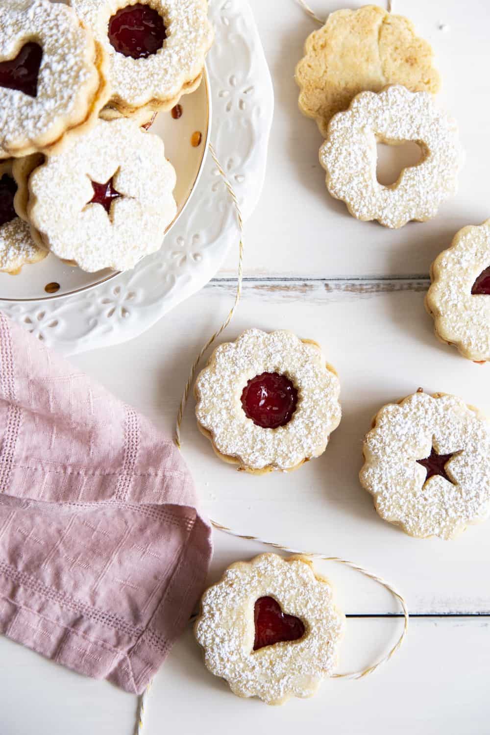 Linzer cookies are a combination of buttery orange infused shortbread and jam, as pleasing to look at as they are to eat. These festive treats are perfect for any holiday or special occasion. Linzer Cookies stacked and laid out in the photo.