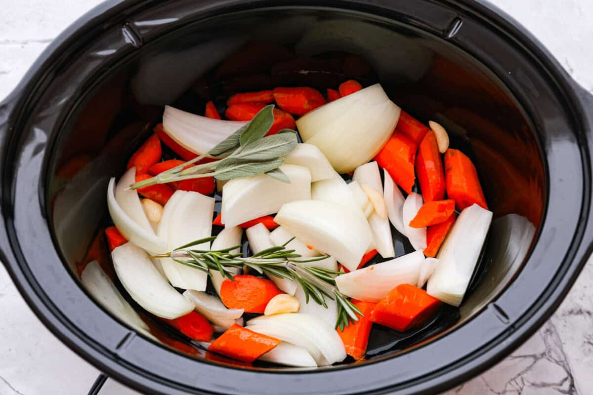 Slow Cooker Turkey Breast is the perfect way to get tender, juicy turkey with a fraction of the prep but all the flavor. Let the slow cooker do ALL the work! Overhead shot of onion, carrots, garlic and sprigs of herbs in the bottom of the crockpot.