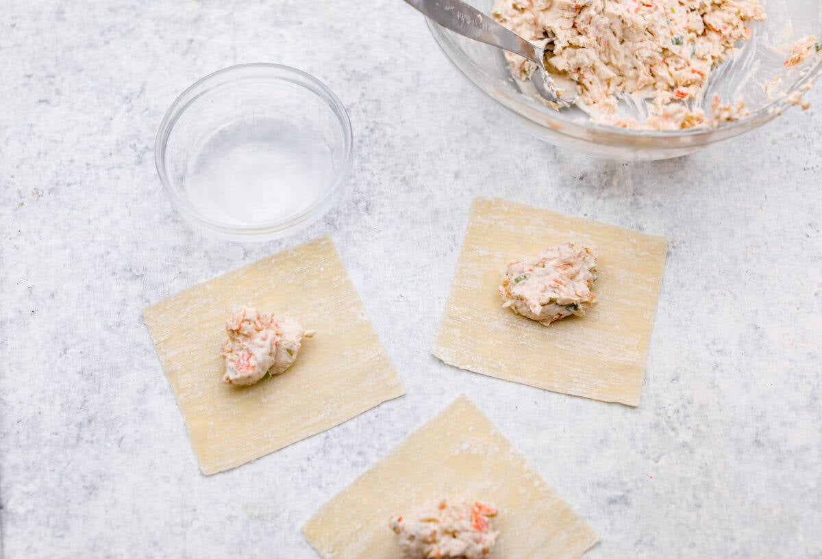 Crab Rangoons are a fun, Asian-inspired appetizer filled with crab meat, smooth cream cheese, and seasoning. Wrapped in a wonton wrapper and deep-fried, they are irresistible! Overhead shot of dollops of mixed filling ingredients on wonton paper.