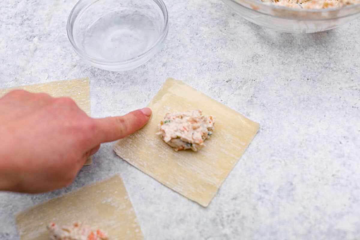 Crab Rangoons are a fun, Asian-inspired appetizer filled with crab meat, smooth cream cheese, and seasoning. Wrapped in a wonton wrapper and deep-fried, they are irresistible! Overhead shot of someone using their finger to wet the edges of the wonton paper.