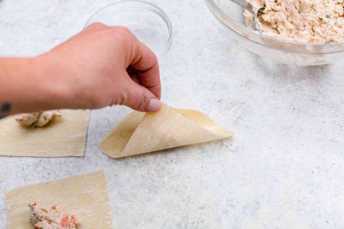 Crab Rangoons are a fun, Asian-inspired appetizer filled with crab meat, smooth cream cheese, and seasoning. Wrapped in a wonton wrapper and deep-fried, they are irresistible! Angle shot of someone pinching two corners of the wonton paper together.