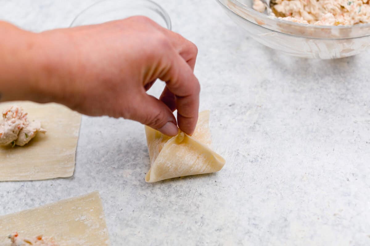 Crab Rangoons are a fun, Asian-inspired appetizer filled with crab meat, smooth cream cheese, and seasoning. Wrapped in a wonton wrapper and deep-fried, they are irresistible! Angle shot of someone pinching all four corners of the wonton paper together with filling inside.