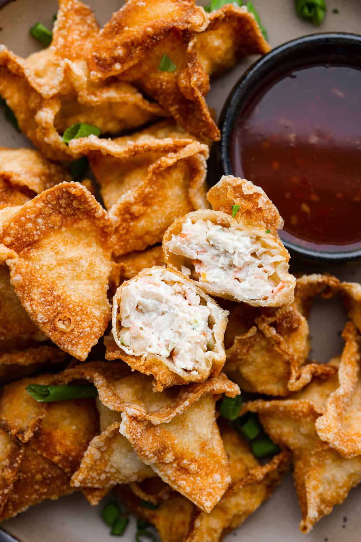 Crab Rangoons are a fun, Asian-inspired appetizer filled with crab meat, smooth cream cheese, and seasoning. Wrapped in a wonton wrapper and deep-fried, they are irresistible! Close up shot of a crab rangoon cut in half over a pile of crab rangoons.