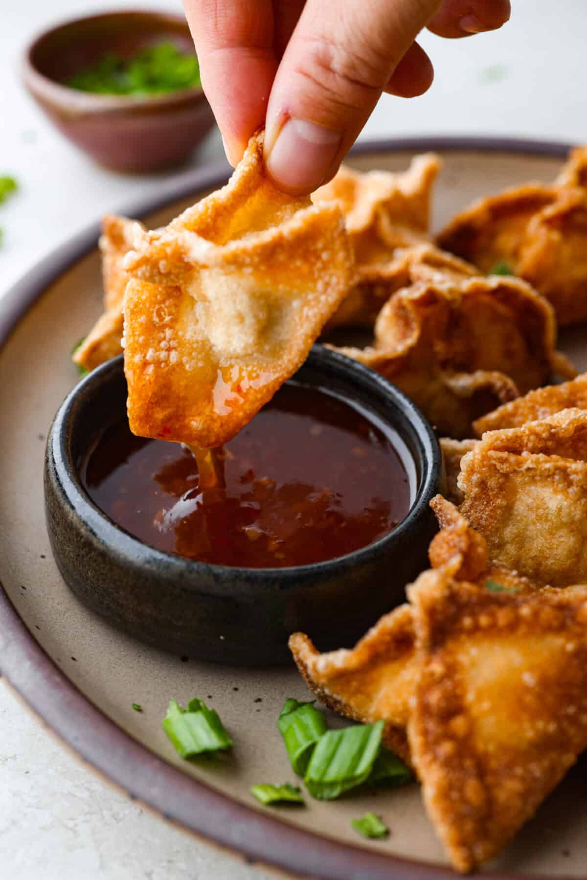 Crab Rangoons are a fun, Asian-inspired appetizer filled with crab meat, smooth cream cheese, and seasoning. Wrapped in a wonton wrapper and deep-fried, they are irresistible! Close up shot of someone dipping their crab rangoon into sauce.