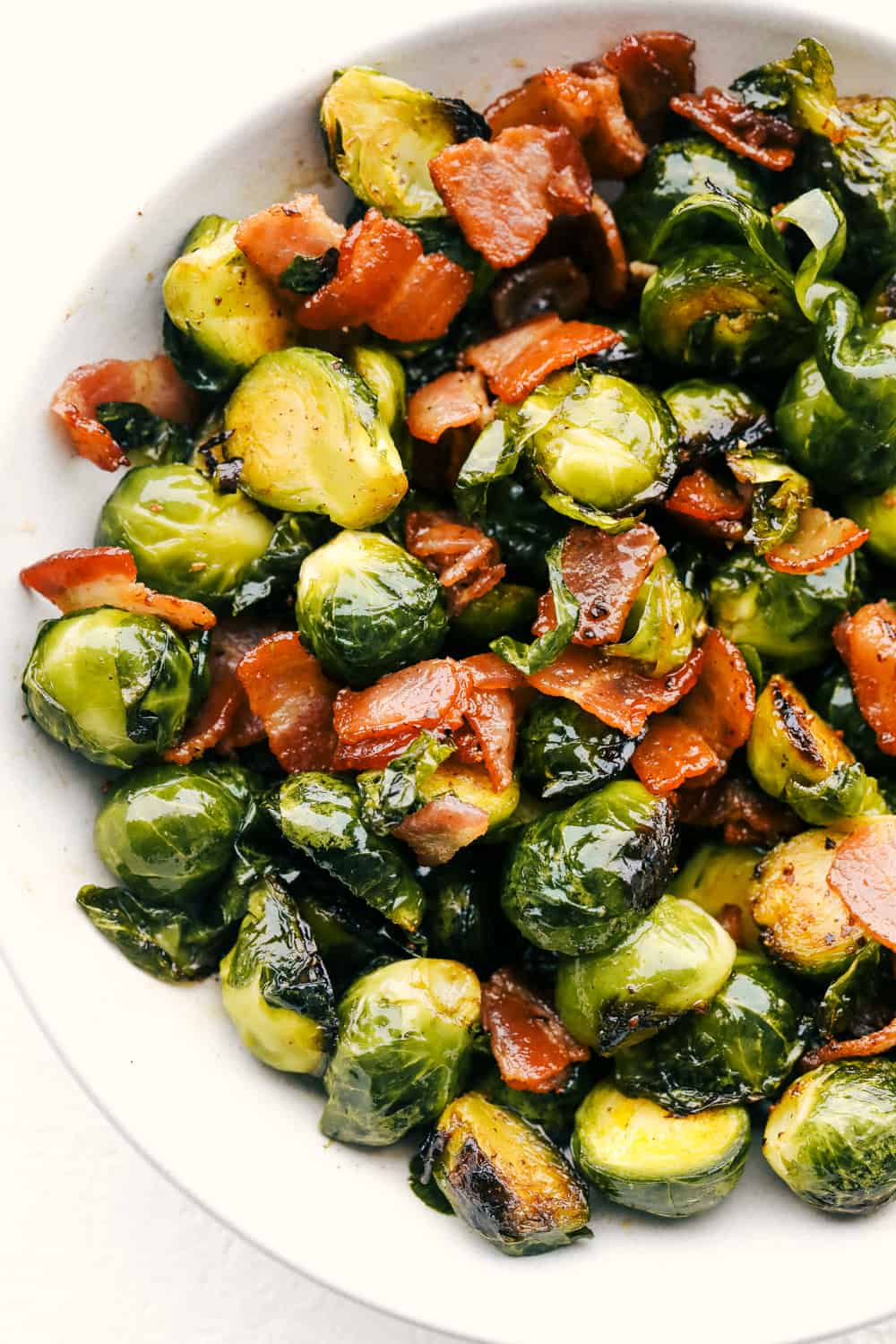 Perfect Sauteed Brussel Sprouts with Bacon is side dish supremacy. Crisp tender brussel sprouts tossed with crispy bacon and a drizzle of maple syrup is pure heaven. Bacon, brussel sprouts tossed with maple syrup on a white plate.