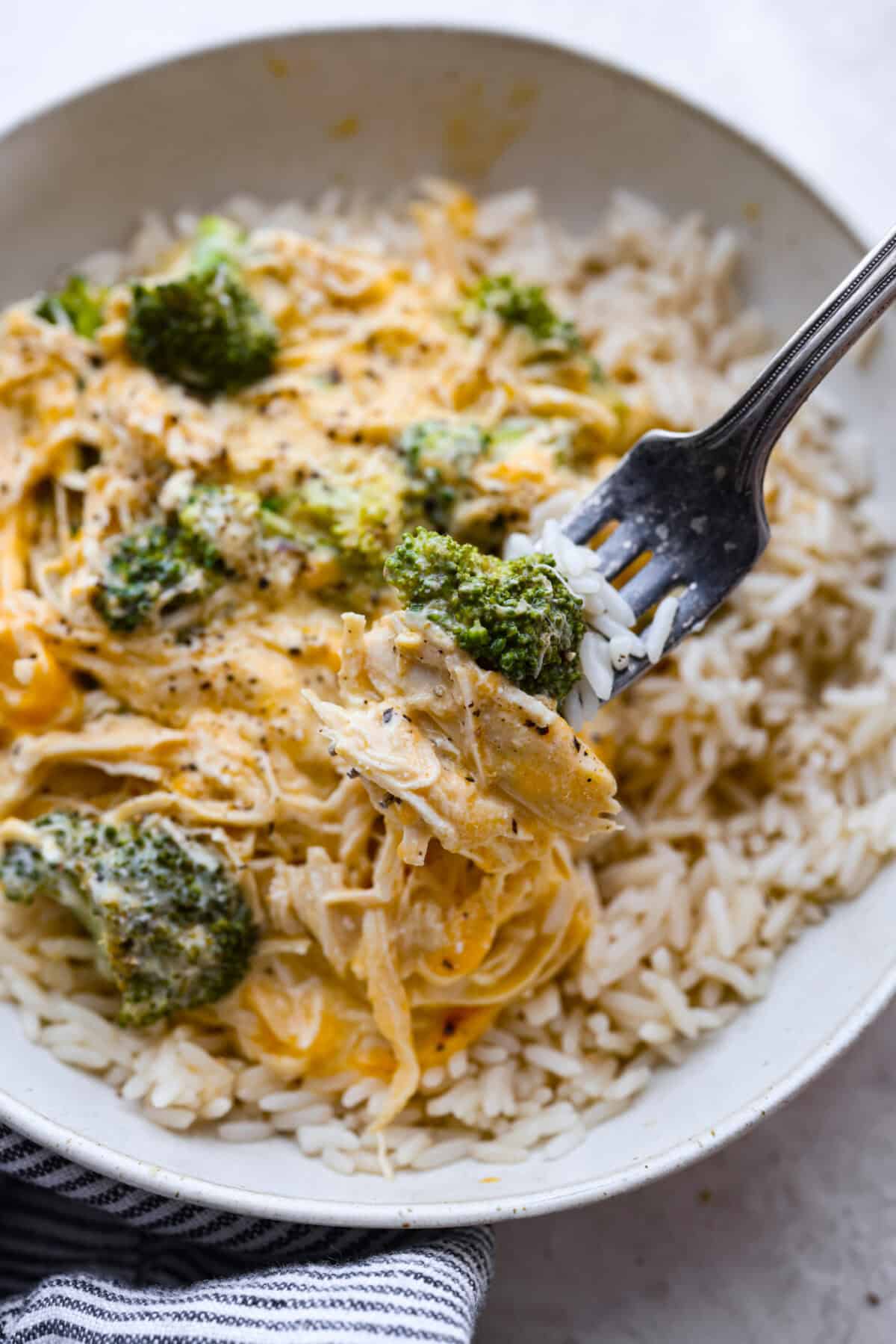 Slow-cooker creamy chicken and broccoli over rice is a super easy, delicious meal the whole family will love! Moist chicken and tender broccoli combine in a cheesy sauce that's absolutely divine.ย Close up shot of a bite of creamy chicken and rice over broccoli on a fork.