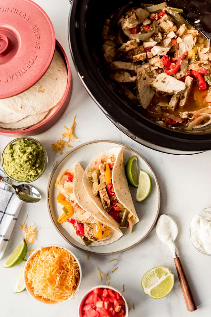 Mexican dinner night is fun and easy with these Crockpot Chicken Fajitas! They are loaded with chicken, bell peppers, onion, and spices, then served with all your favorite toppings. Chicken fajitas with toppings next to a slow cooker.