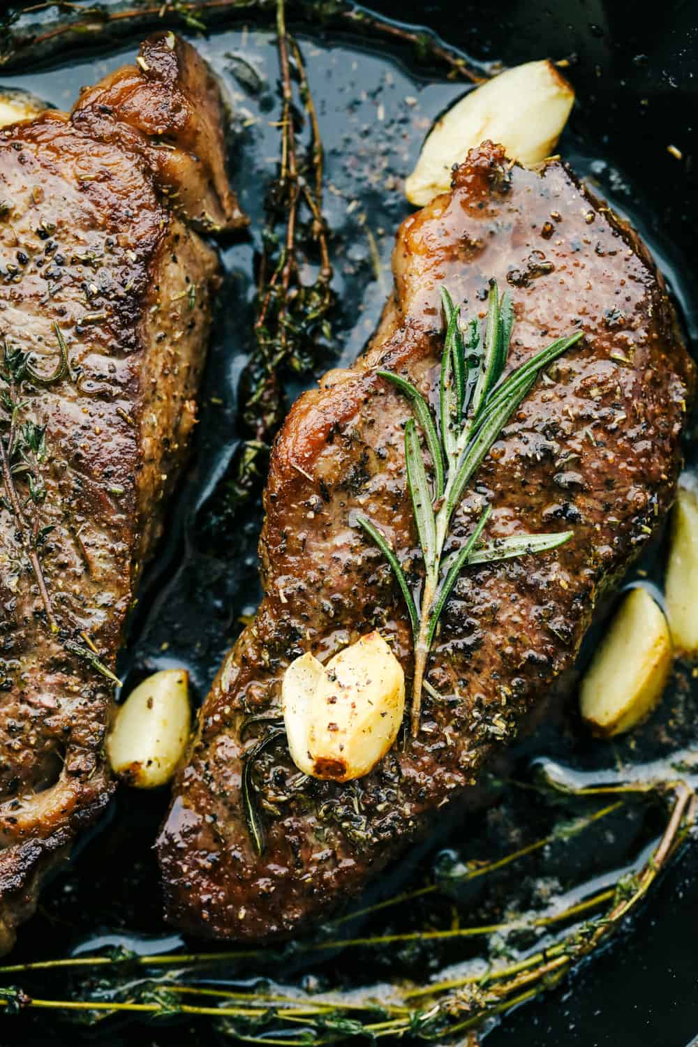 Skillet seared New York Steak is one of the best steaks you'll ever have!ย It's tender, juicy and full of flavor! Two steaks in a black pan with garlic over top.