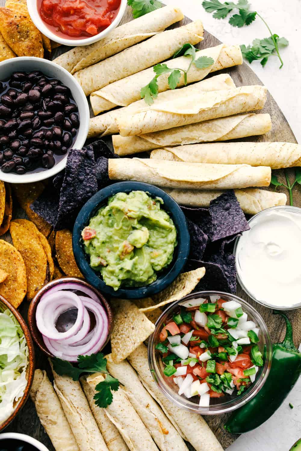 This taco board is everything you need to have for the ultimate get together! It is the perfect combination! Taquitos on a board with bowls of dips, vegetables and chips all around.