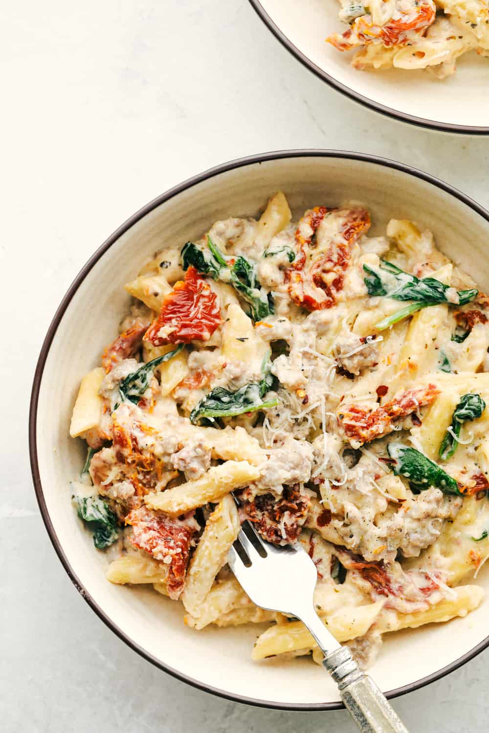 Creamy Tuscan sausage pasta is pretty much a one pan wonder you will want to cook over and over again!ย Perfect for a busy night, this is a hearty comforting dish that will truly satisfy. Tuscan sausage pasta in a white bowl with a fork.