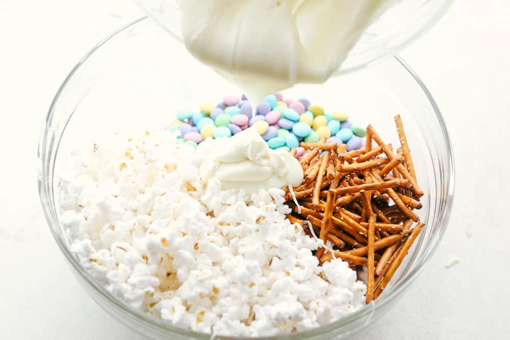 Funfetti Popcorn or Bunny Bait is a fun springtime treat that is sure to satisfy! This is a sweet salty colorful treat perfect for Easter or any other special occasion! Pouring melted white chocolate over popcorn, M&Ms and pretzels.