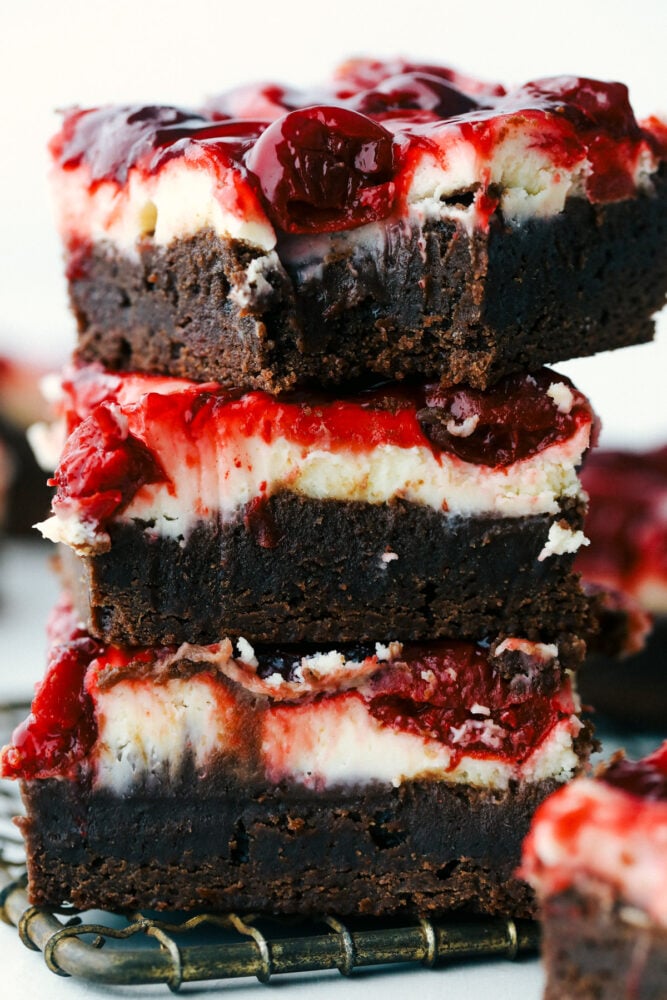 Cherry cheesecake brownies are fudgy brownies with cheesecake and premium cherry pie filling swirled on top! These brownies are rich and creamy and have three delicious desserts in one! Brownies stacked on top of each other and one with a bite out of it.