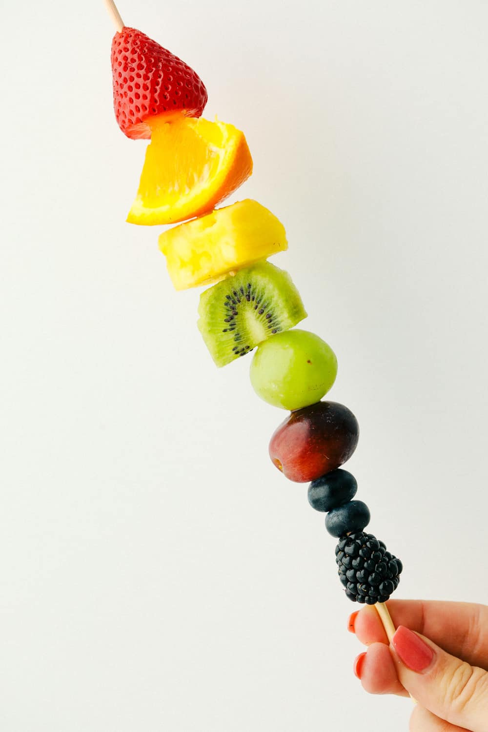 Fresh Rainbow Fruit Skewers are a super easy and fun sweet treat. Fruit skewers are a colorful, fun and yummy way to eat the recommended servings of fruit! Holding a rainbow kebab.