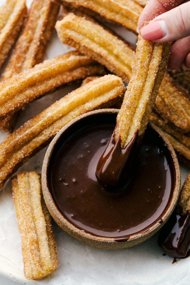 All I have to hear is the word, Churros, and my mouth starts to water. Light fluffy dough is fried and then covered in cinnamon and sugar, totally heavenly! Holding a churro with chocolate sauce on it, with fired churros all around.