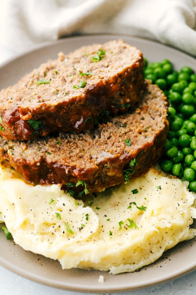 Instant Pot Meatloaf is the best meatloaf I have ever had! Full of vegetables and flavor, it comes out perfectly cooked, tender and moist every time! Sliced meatloaf on a plate with a side of peas and mashed potatoes.