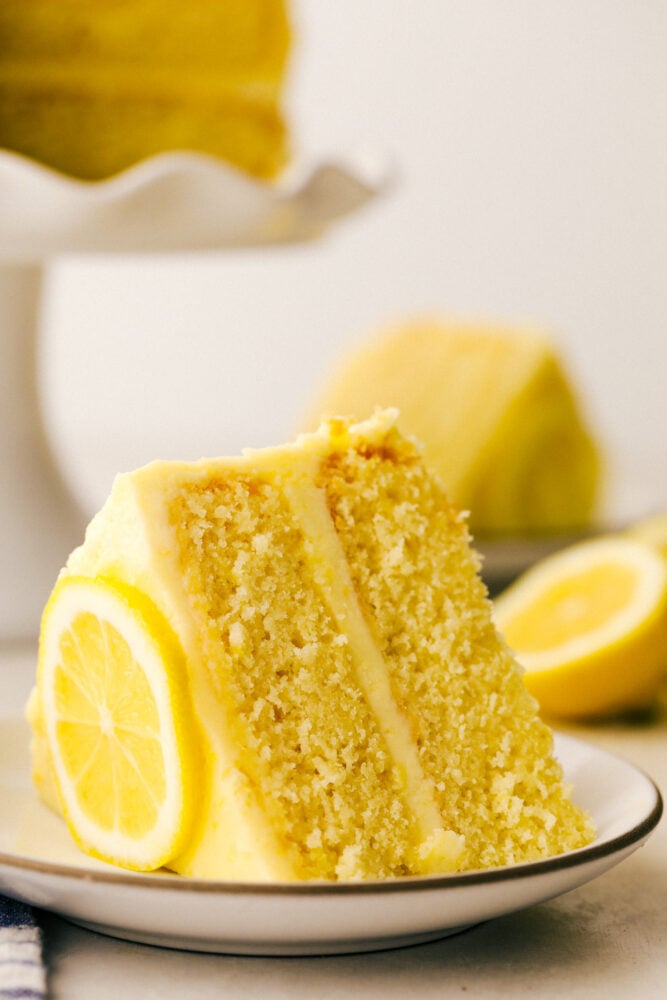Lemon Cake is everything you want in a sweet, yet zingy cake! Tender moist layers of refreshing lemon cake are smothered with a tart but sweet frosting. A slice of cake with a lemon garnish on a plate.