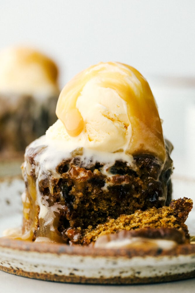 Sticky Toffee Pudding is an English dessert that you are going to absolutely love! This moist sponge cake contains finely chopped dates, a rich toffee sauce, and served with vanilla ice cream. A bite taken out of the cake with ice cream and sauce dripping down.