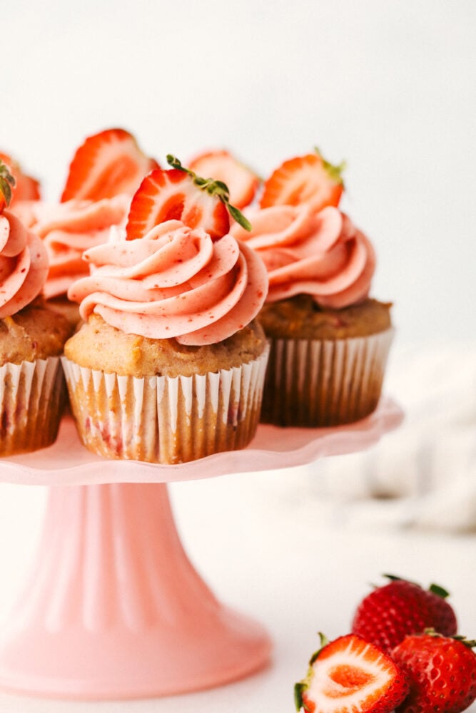 Strawberry Cupcakes are made with fresh and freeze-dried strawberries to create an incredible burst of flavor. Pretty and pink, these cupcakes are sure to be the perfect treat. Strawberry cupcakes on a platter with frosting and strawberries on top.