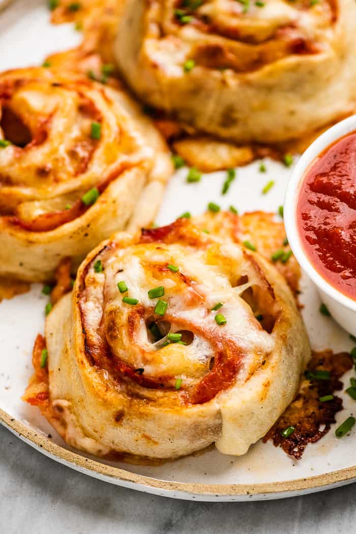 Pizza pinwheels are a super easy, family-friendly lunch or dinner. They're made with soft pizza dough and your favorite toppings rolled up then sliced into rounds. Once they're baked, they become cheesy and delicious bites that your whole family will love. A closeup of pizza pinwheels on a plate with dipping sauce.