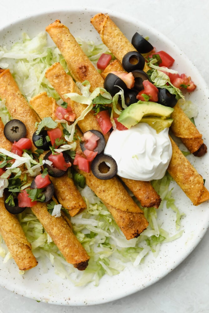 These air fryer frozen taquitos are quick and easy to make for a snack, dinner or side dish! Inside each taquito is packed with protein and flavor! Air fryer taquitos on a white plate with sour cream, olives, tomatoes and lettuce.