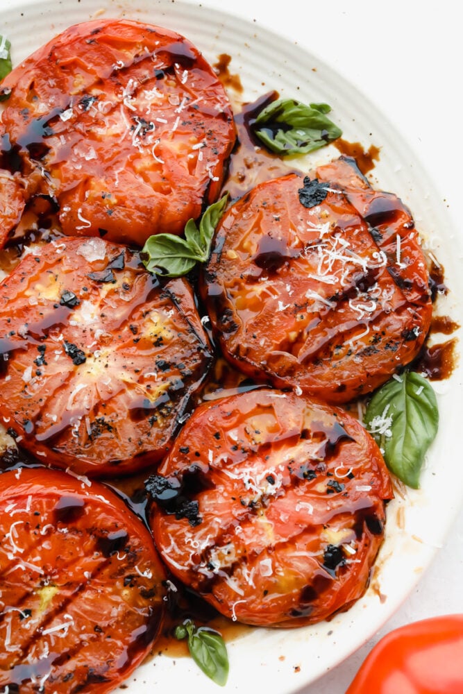 Super easy and super delicious, you are going to love these juicy Grilled Tomatoes! Each slice is cooked to perfection and bursting with luscious, savory flavors in every bite. Grilled tomatoes on plate with balsamic vinegar glaze.