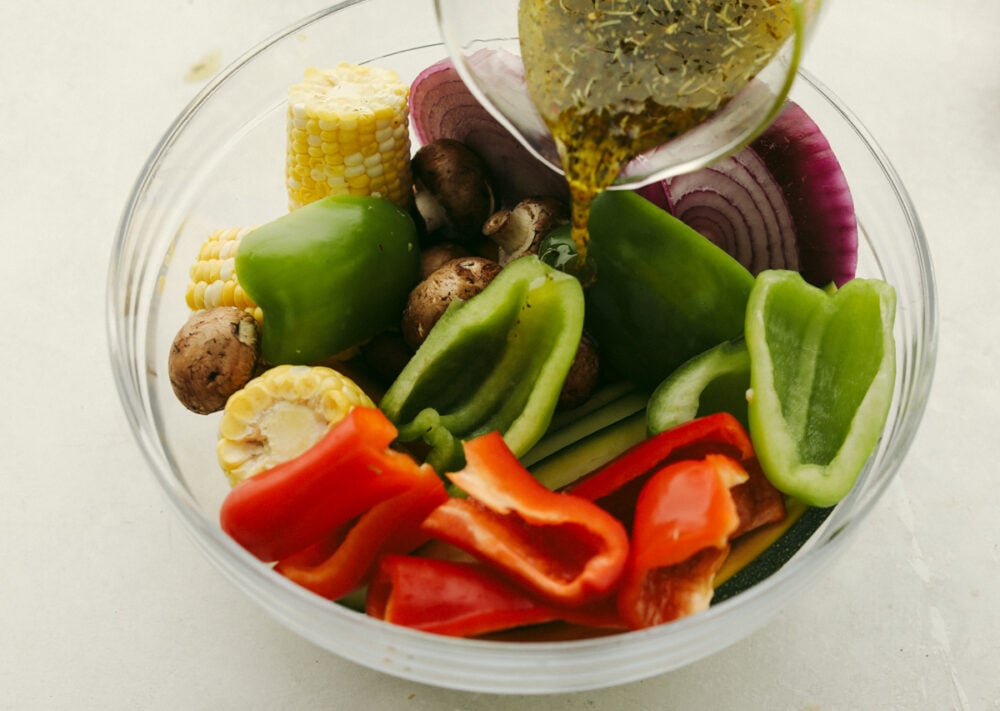 These delicious Grilled Vegetables are the perfect side to enhance any meal. They are drizzled with a savory garlic marinade that amplifies each vegetable's amazing flavor and texture! Mixed vegetables in bowl with marinade being poured over it.