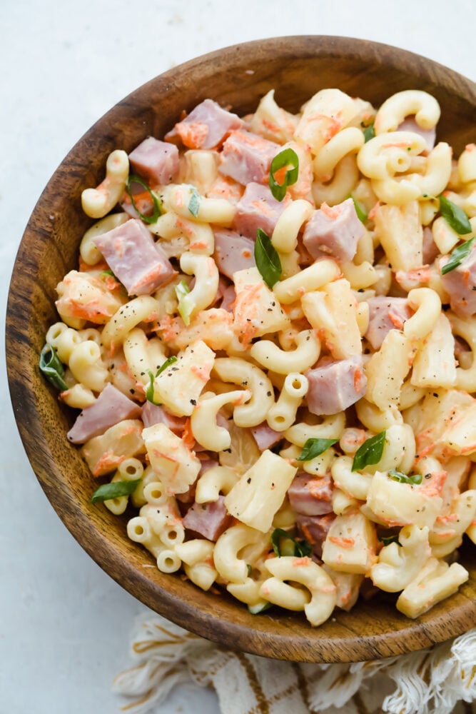 Hawaiian Macaroni Salad is the perfect side dish! It is a delicious macaroni pasta salad with ham, pineapple, shredded carrots, and green onions, and a tangy dressing. A wooden bowl with hawaiian macaroni salad in it.