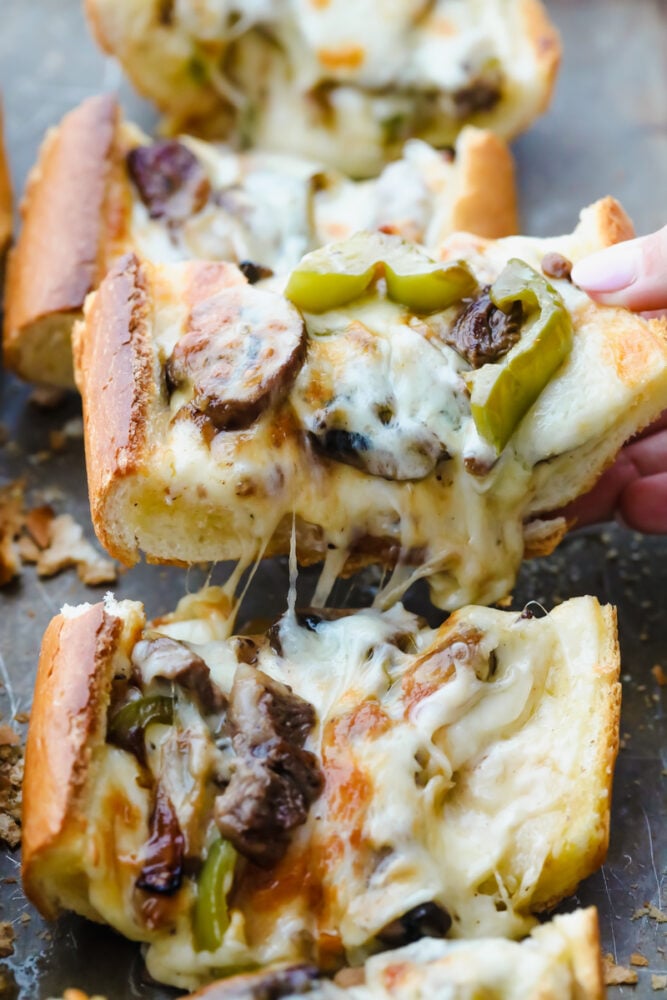 Philly Cheese Steak Cheesy Bread with just a few simple ingredients is the taste of Philly for a crowd! It comes together in just minutes with major flavor in each bite! Piece of Philly cheesesteak cheesy bread being pulled from pan