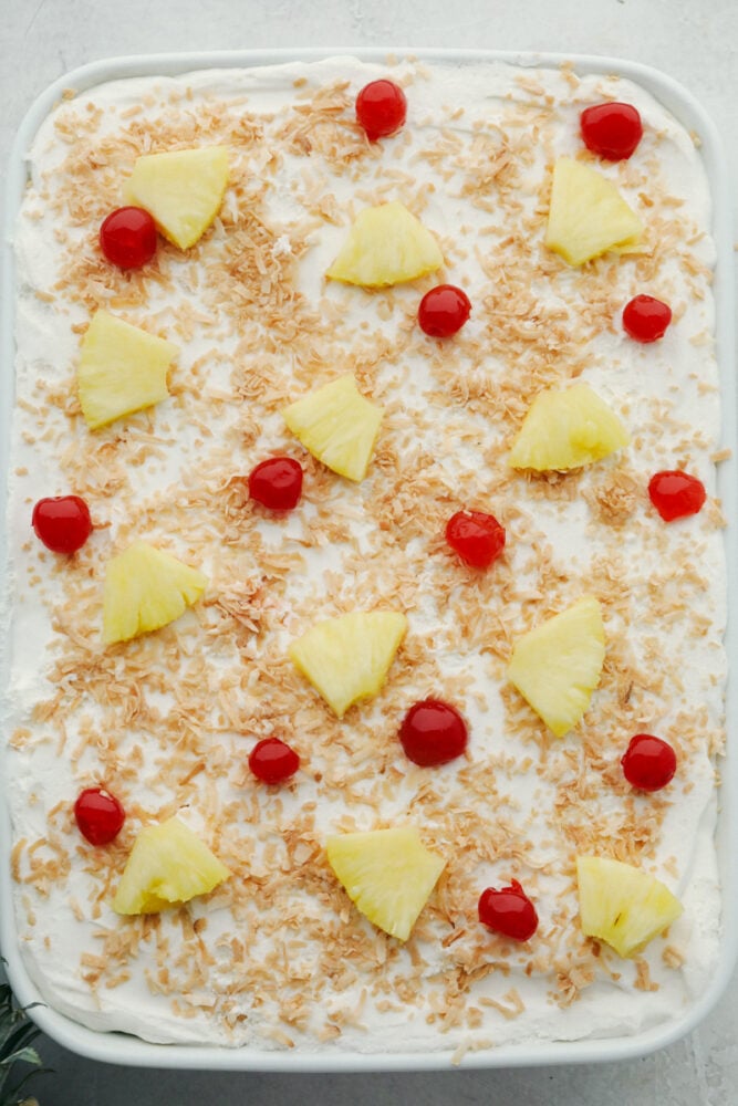 Pina Colada Poke Cake is a little slice of tropical paradise on a plate! Rich in flavor this is a light fluffy cake everyone is going to love! Decorated pina colada poke cake with pineapple, and maraschino cherries.