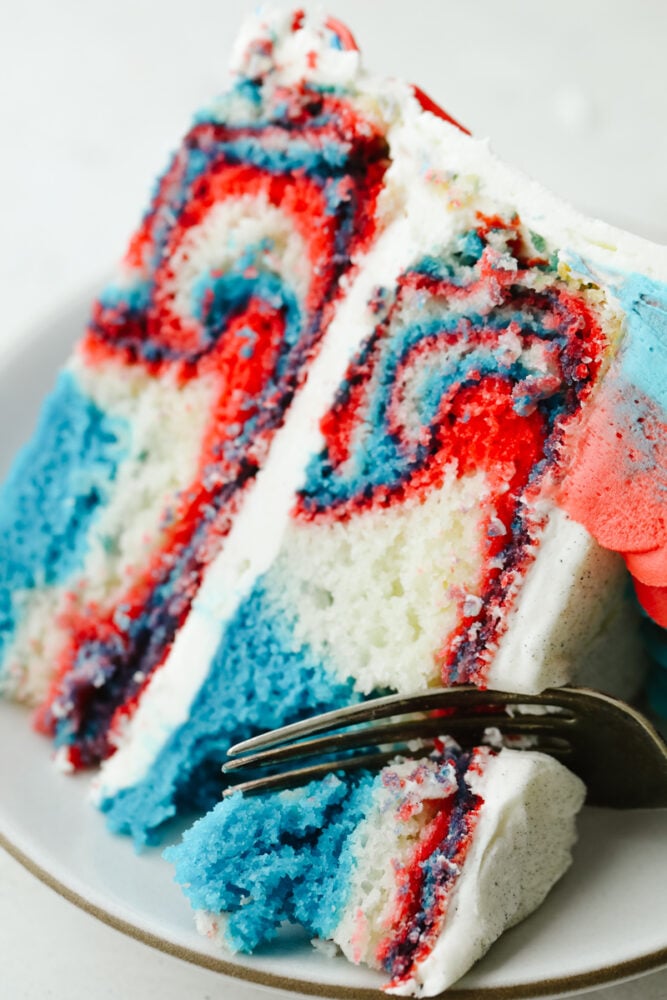This Red, White and Blue Cake is going to steal the show for your 4th of July celebrations! Tender moist cake is swirled in the sweetest tie-dye pattern! Cutting into the cake with a fork.