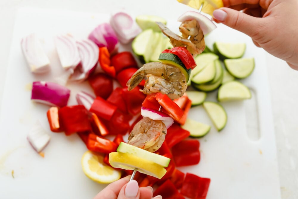 Shrimp Kabobs are marinated and grilled to perfection for the most scrumptious summer meal. Full of zesty flavors the whole family is going to love these kabobs! Placing the shrimp and vegetables onto skewers.
