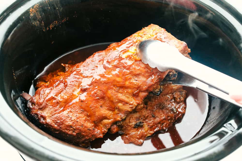 Myย Slow Cooker Ribsย are hands-down one of my favorite recipes on the site! They fall off the bone, are coated in sticky-sweet sauce, and are unbelievably easy to make. Someone taking cooked ribs out of the slow cooker with tongs.