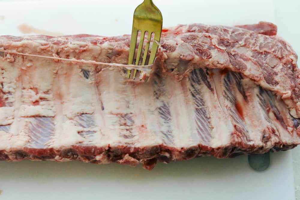 Myย Slow Cooker Ribsย are hands-down one of my favorite recipes on the site! They fall off the bone, are coated in sticky-sweet sauce, and are unbelievably easy to make. Rib rack with someone using a fork to take the membrane of the back.