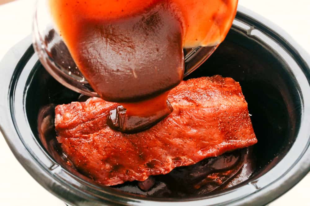 Myย Slow Cooker Ribsย are hands-down one of my favorite recipes on the site! They fall off the bone, are coated in sticky-sweet sauce, and are unbelievably easy to make. Someone pouring bbq sauce over seasoned ribs in the crockpot.