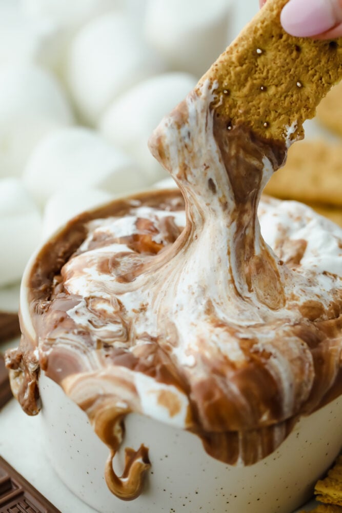 S'mores Dip is an easy way to get all the ooey gooey goodness from a s'more without the roasting! This is such a fun sweet treat that everyone can enjoy without building a hot fire A graham cracker dipping into the chocolate and marshmallow.