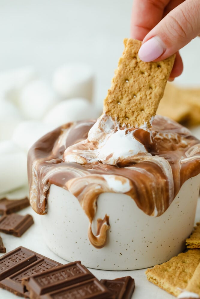 S'mores Dip is an easy way to get all the ooey gooey goodness from a s'more without the roasting! This is such a fun sweet treat that everyone can enjoy without building a hot fire A graham cracker dipping into s'more dip.