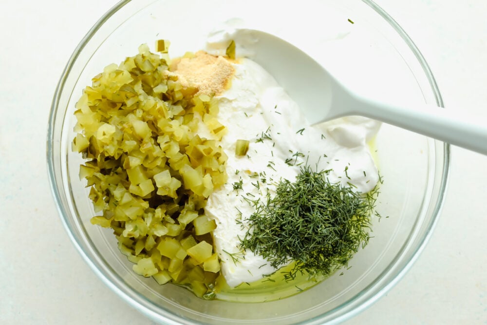 Calling all pickle lovers! Loaded with chopped pickles and herbs, dill pickle dip is bursting with flavor and absolutely irresistible! Dill pickle dip ingredients in a glass bowl.