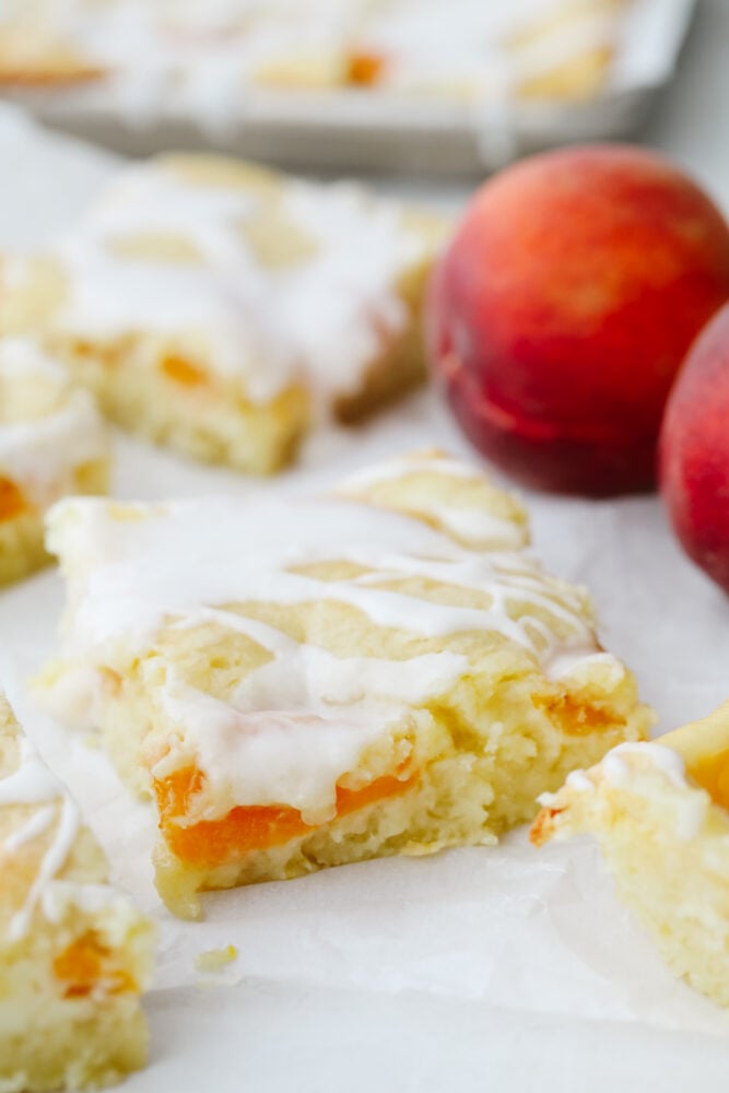 Peaches and Cream Pie Bars are delicious bars that have a creamy cheesecake and peach pie filling in the center. They are drizzled in a glaze and taste just like you are biting into a peach pie! Peaches and cream pie bars on parchment paper.