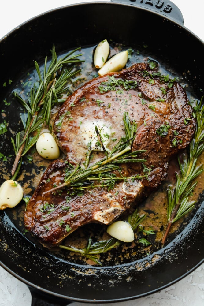Achieving steakhouse-worthy flavors doesnโt have to be difficult! The ultimate ribeye steak is juicy, flavorful, and seared to perfection all in the comfort of your kitchen. Cooked ribeye steak in a skillet, garnished with garlic, rosemary, and thyme.