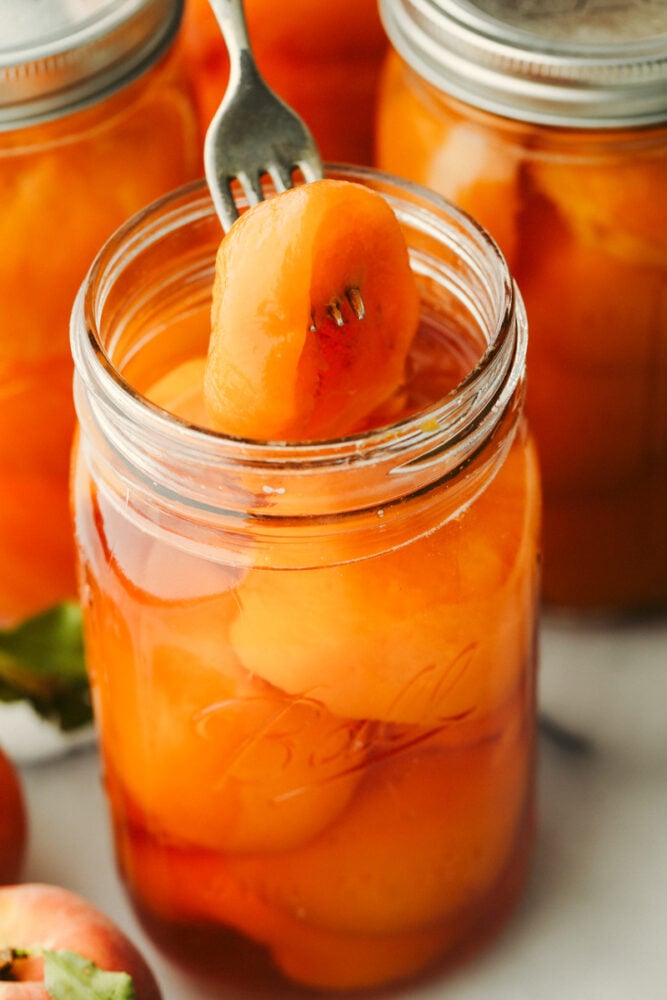This in-depth guide to canning peaches gives you everything you need to know to have delicious peaches on hand year-round! It is easier than you would think and yields the sweetest, most delicious results each time. Closeup of canned peaches with a peach skewered on a fork.